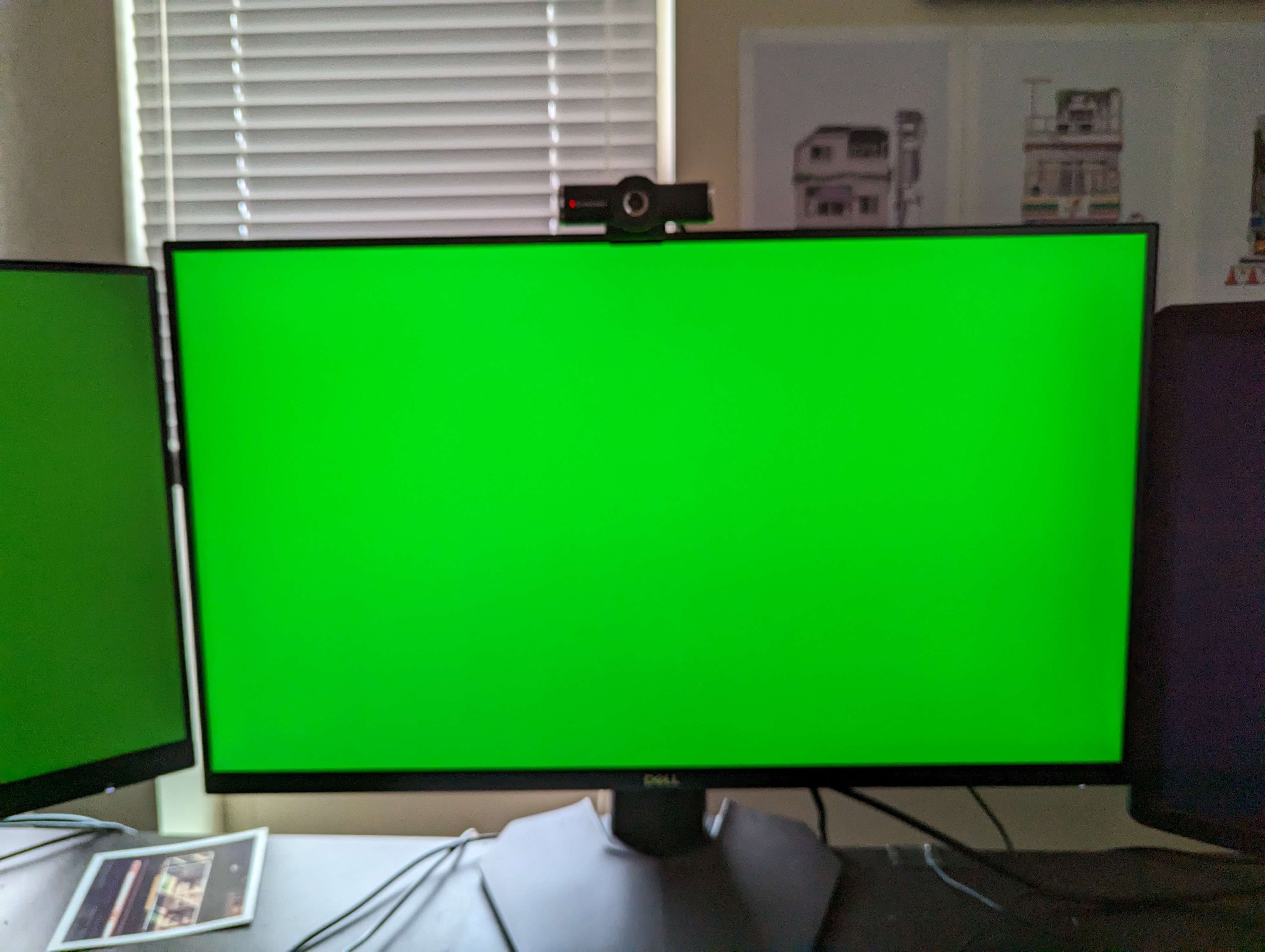 Photo of a computer with three monitors.  Two of the monitors are entirely green, and the rightmost monitor is black.  The monitors are on a black desk, there’s a window with the blinds closed behind it, and there are some art prints on the wall along with one on the desk.