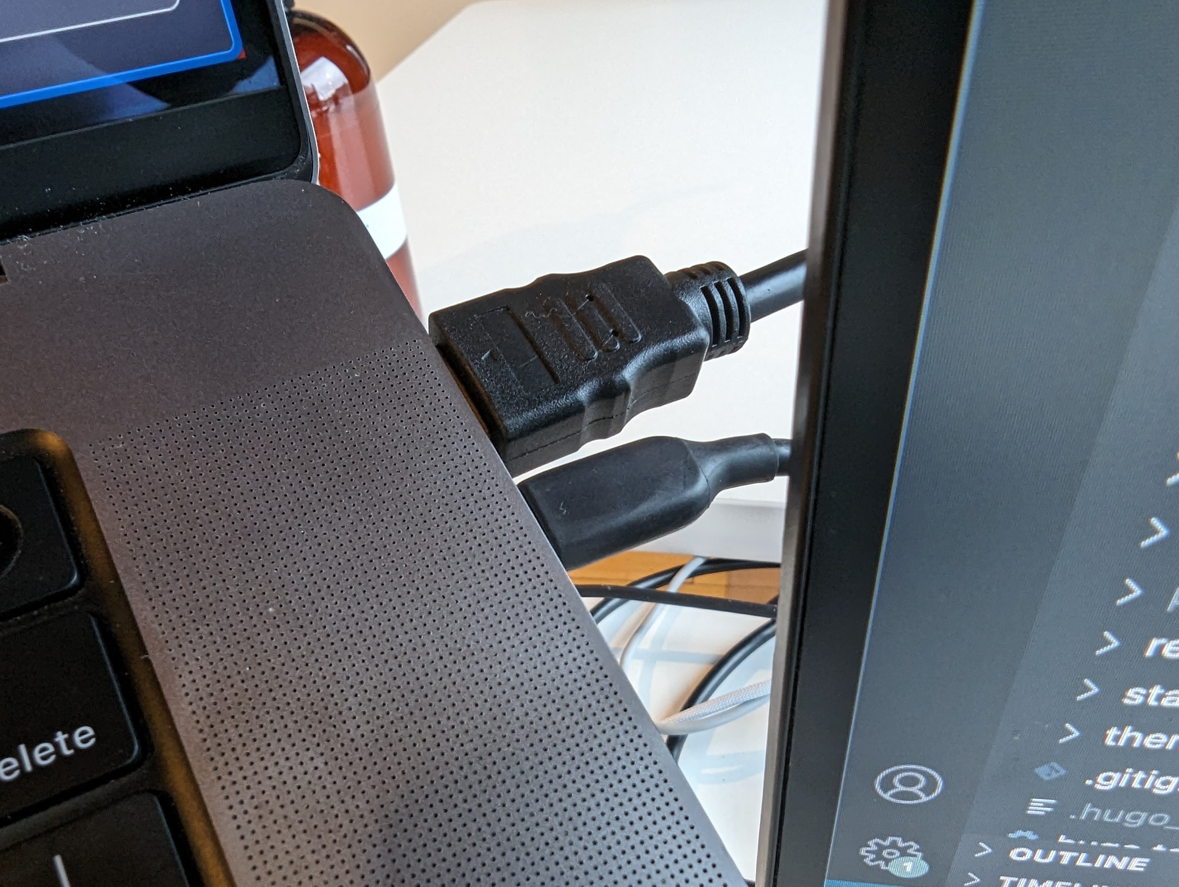 A close-up photograph of a black USB-C cable and a black HDMI cable plugged into the upper right side of an M1 Macbook Pro side by side.  There is a small sliver of monitor with the file tree of VS Code open visible on the right side.